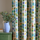 A close-up of patterned curtains hanging in a room, featuring a vertical stripe design with irregular watercolor-like dots in green, blue, yellow, and purple on a cream background. A light blue ceramic pitcher with a wooden handle and green eucalyptus stems sits on a distressed green cabinet to the left.